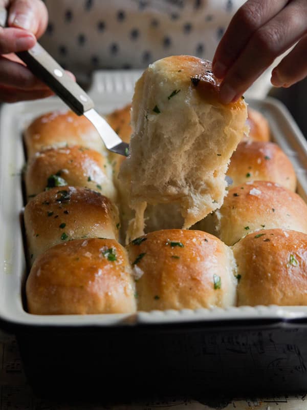 A hand is pulling a fluffy, golden brown dinner roll garnished with herbs from a baking dish using a knife. The rolls are shiny and appear freshly baked with a soft texture.